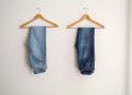 Two pairs of blue denim jeans neatly hanging on wooden hangers against a plain wall.