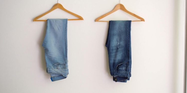 Two pairs of blue denim jeans neatly hanging on wooden hangers against a plain wall.