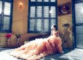 A bride in a luxurious gown sits gracefully in a sunlit room, exuding elegance.