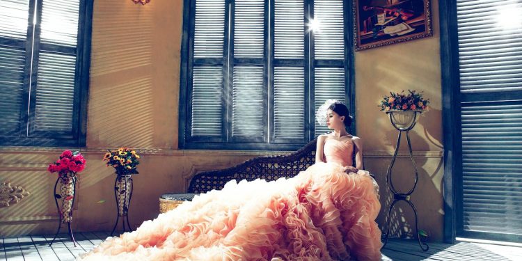 A bride in a luxurious gown sits gracefully in a sunlit room, exuding elegance.