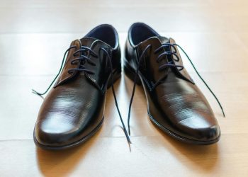 A pair of shiny, elegant black leather dress shoes with laces on a wooden floor.