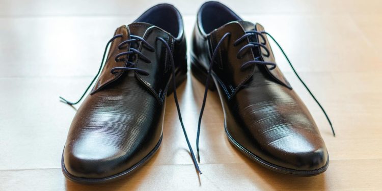 A pair of shiny, elegant black leather dress shoes with laces on a wooden floor.
