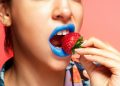 Close-up portrait of a woman with blue lipstick holding a fresh strawberry.