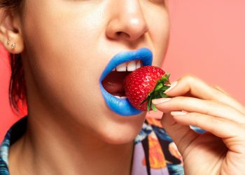 Close-up portrait of a woman with blue lipstick holding a fresh strawberry.