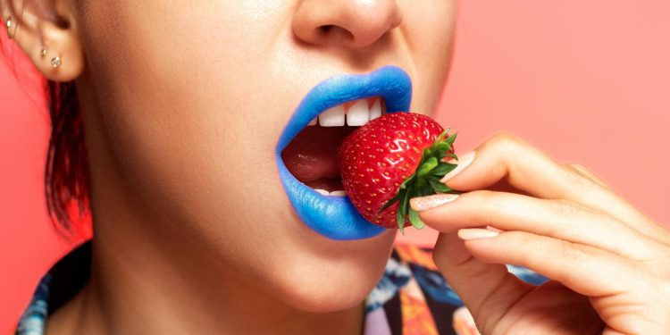 Close-up portrait of a woman with blue lipstick holding a fresh strawberry.