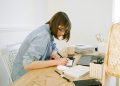 A young woman working from home, writing notes at a desk with a laptop and papers.