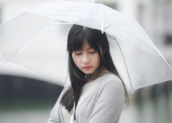 Asian woman with a transparent umbrella stands outdoors during a rainy day, embodying a serene and contemplative mood.