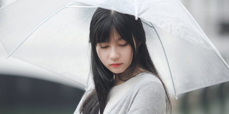 Asian woman with a transparent umbrella stands outdoors during a rainy day, embodying a serene and contemplative mood.